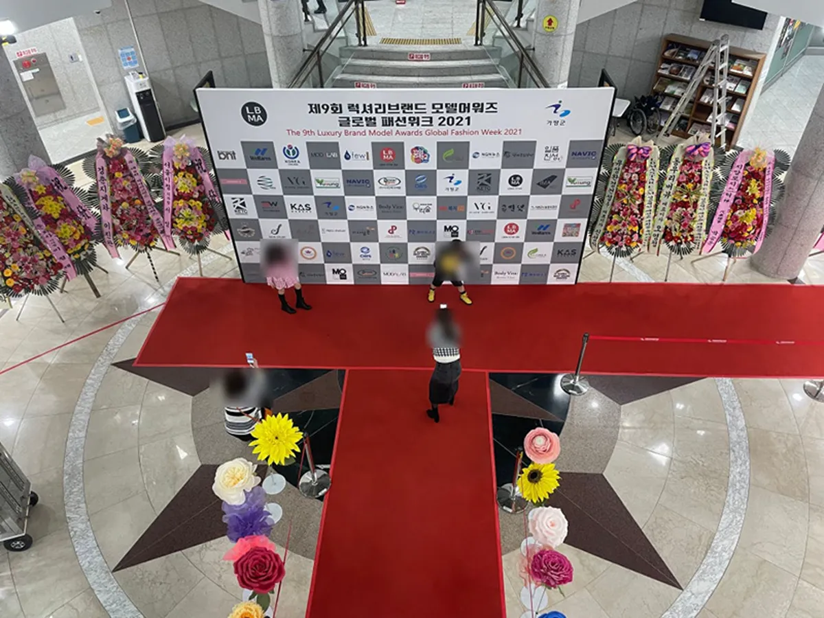 실내 행사장 가운데 레드카펫 T자 무대와 백월, 화환이 놓인 기념식 세팅을 위에서 본 모습